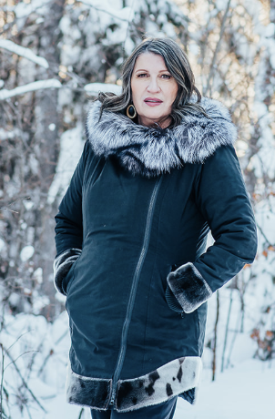 Woman standing in a snowy forest wearing a dark winter coat with fur trim on the hood, cuffs, and hem. She has long, wavy dark hair and is looking at the camera with a neutral expression. Snow-covered trees are visible in the background.